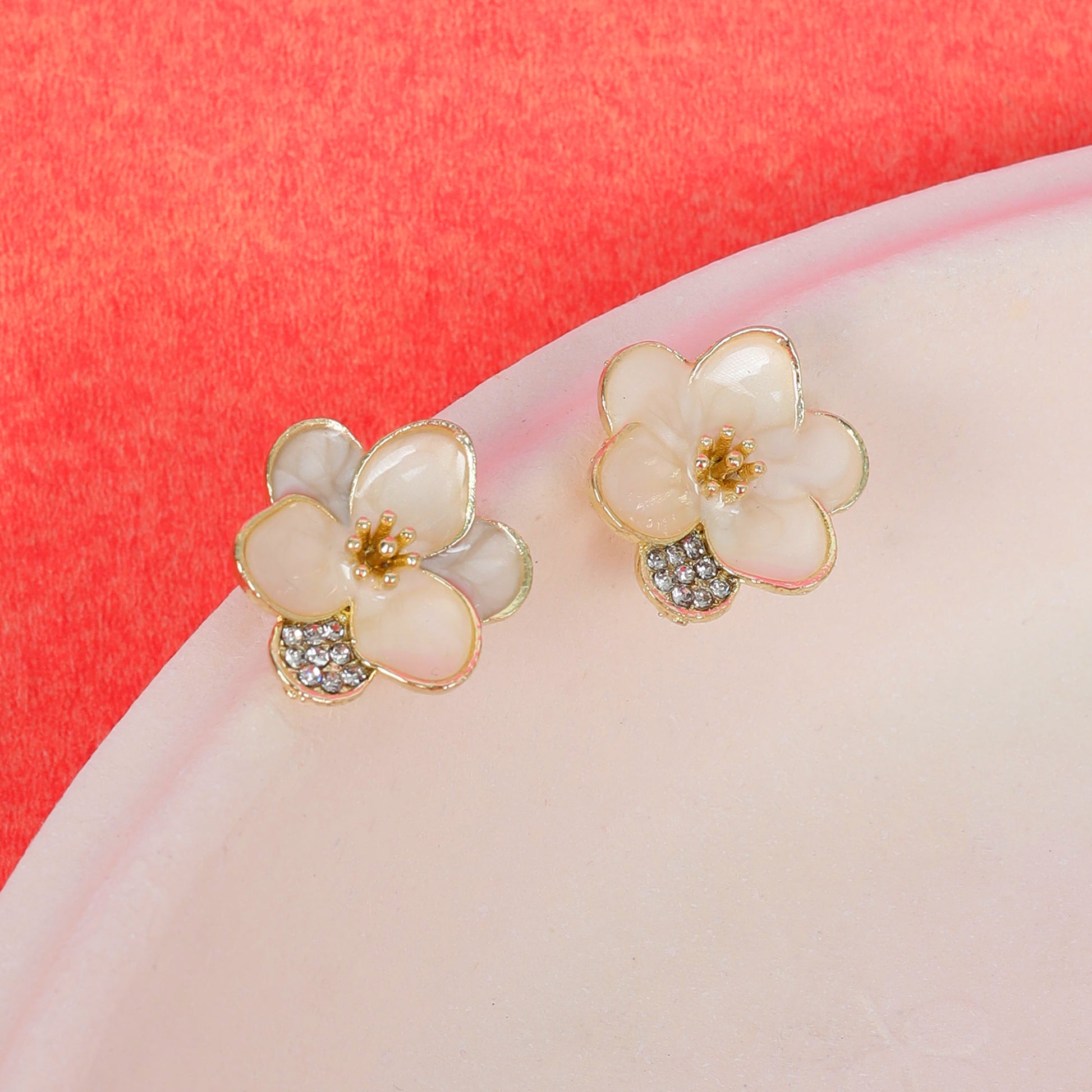 A pair of Enamel flower earrings with gold trim, cream-colored petals, and crystal accents, displayed on a round pale surface against a red background.